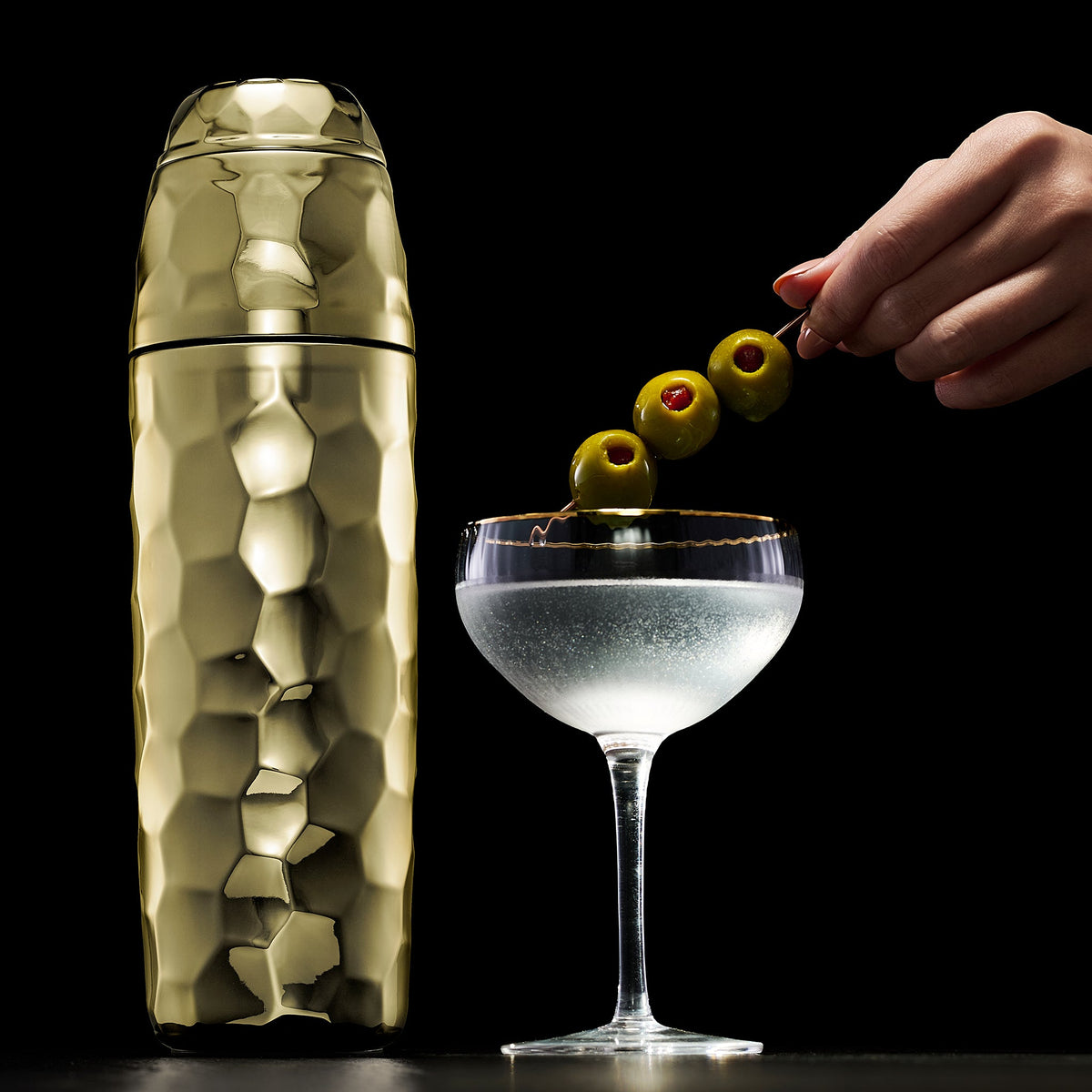 Gold hexclad cocktail shaker next to a cocktail made with olives on a black background